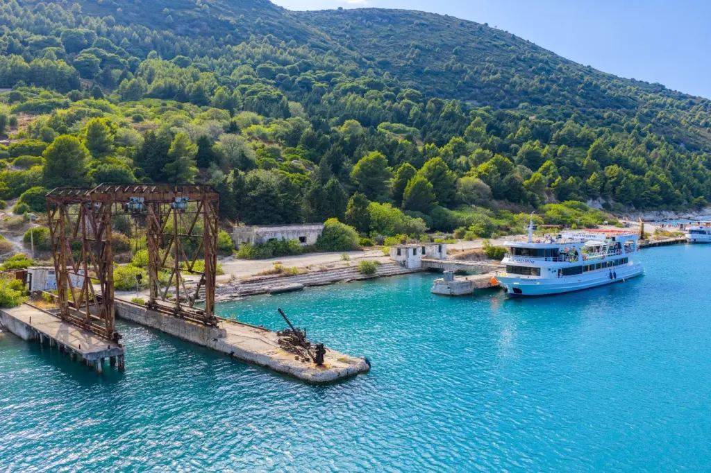 A rusted gantry crane on an old concrete quay, a white passenger ship moored at the pier and pine-covered hills behind